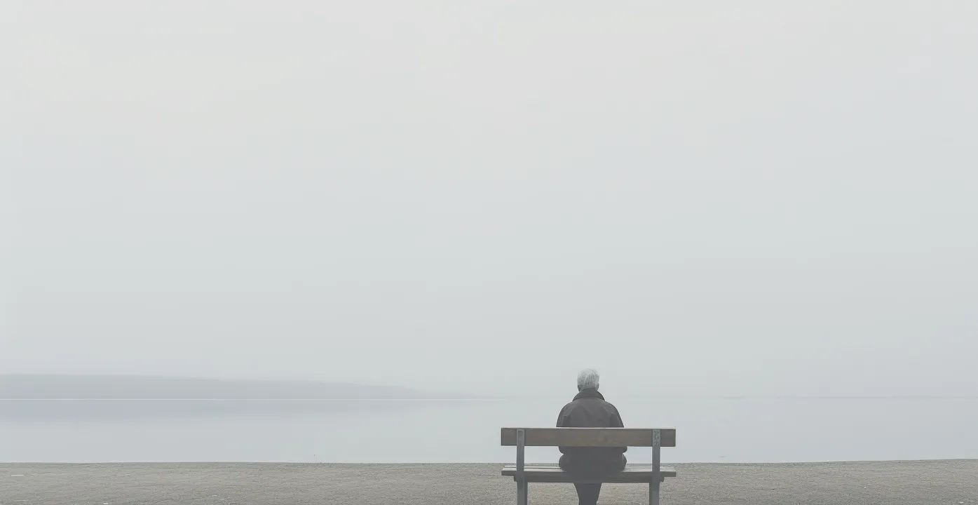 Wide misty seascape with an elderly figure sitting on a bench, seen from behind.